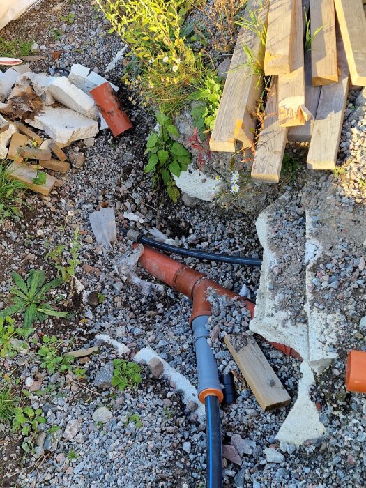 Grävt område med synliga vattenrör och avloppsrör, omgivna av grus och plankor. Byggmaterial och växter syns också.