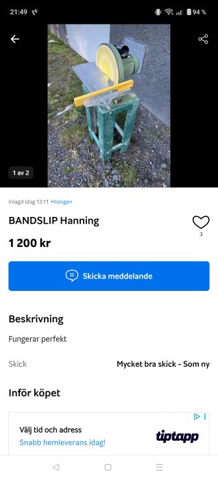 Verktygsbild av en bandslipmaskin på ett grönt metallstativ, placerad utomhus på grus och gräs, bredvid en grå vägg.
