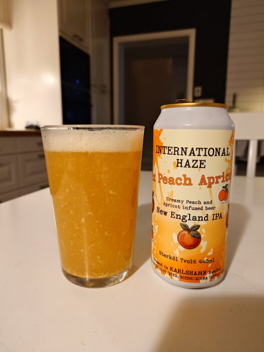 Glas med skummande öl bredvid en burk av "International Haze Peach Apricot New England IPA" på ett köksbord.