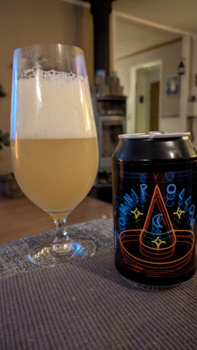 Ett glas med skummande öl bredvid en Omnipollo-ölburk med färgglatt hattmönster, i ett hemtrevligt rum med kamin i bakgrunden.