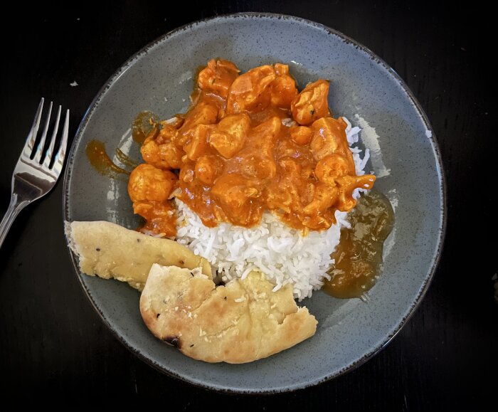 Tallrik med kyckling tikka masala, ris, naanbröd med vitlökssmör och mango chutney, serverad på ett mörkt bord med en gaffel.