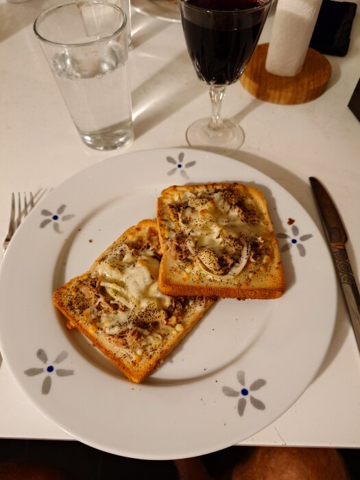 Två rostade bröd med tonfisk, lök och smält ost på en blomdekorerad tallrik, bredvid ett glas rött vin och ett glas vatten.