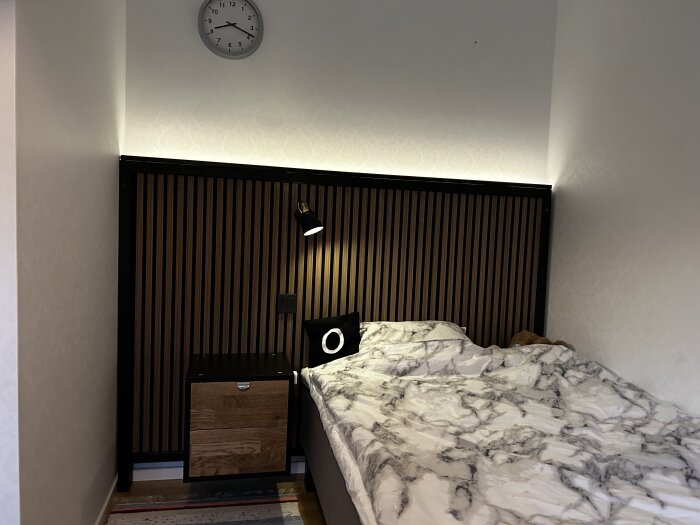 Säng med marmormönstrat överkast, väggmonterad svart lampa och nattduksbord i trä med provisoriskt handtag, mot akustikpanel i valnötsfärg.