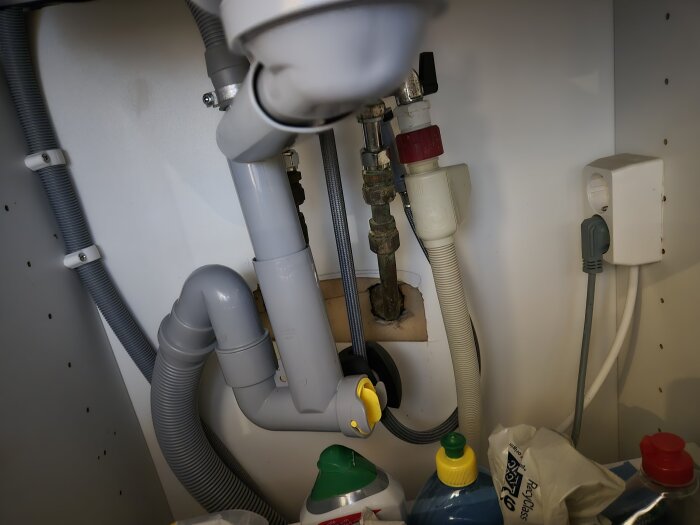 Rör och avloppssystem under diskbänk med gråa och vita slangar samt ventil, omgiven av rengöringsprodukter.