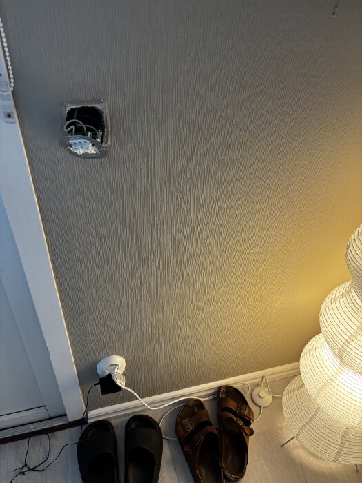 Vägg med en öppen eluttagspanel och exponerade kablar, en lampa och skor på golvet bredvid.