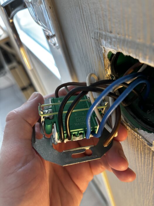 Elektrisk kopplingsbox med flera trådar, hålls i handen, synlig vägguttag och strömbrytare i bakgrunden. Kopplingsproblem diskuterat i texten.