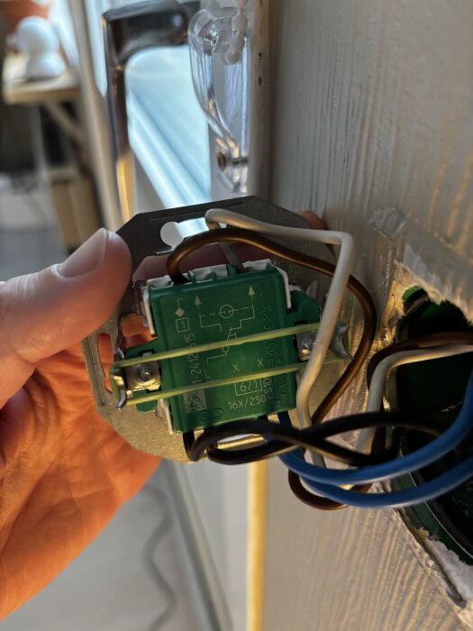En hand håller en strömbrytare med flera kopplade kablar som sitter i en vägg, med närbild på kopplingsdetaljer och färgade ledningar.