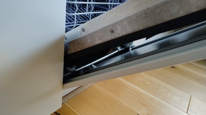 Diskmaskin med lucka halvöppen, synligt justeringshjul, och övre korg delvis gömd. Träklossar limmade under för att centrera luckan. Bild tagen underifrån.