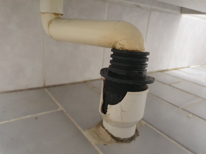 Plastavloppsrör under handfat, svart gummipackning, spricka synlig vid övergången mot golvet, del av äldre badrumsinstallation.