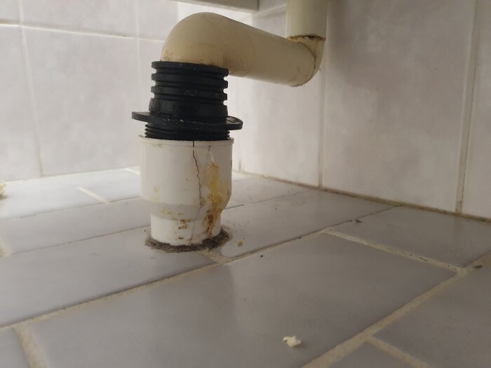 Sprucket avloppsrör i plast under badrumskommod, med synlig spricka och skarvning vid gummitopp. Rördel ligger mot vitkaklat golv.