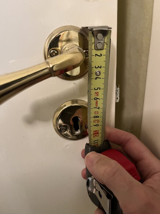 Person mäter avståndet från dörrhandtag till nyckelhål på en toalettdörr med måttband.