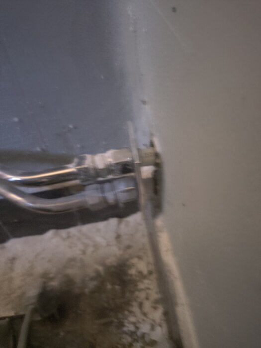 Vattenrör i en vägggenomföring, rördelar synliga. Vy mot betongvägg, rör anslutet i koppling, antydan till slitage eller smuts på väggen.