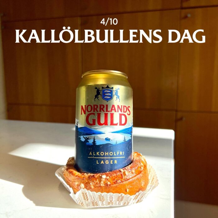 Alkoholfri lagerburk "Norrlands Guld" placerad på en kanelbulle med text "Kallölbullens dag 4/10" i bakgrunden.