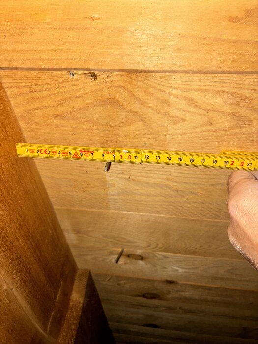 Hand håller ett måttband vid trätak, visar avstånd från takstolar.