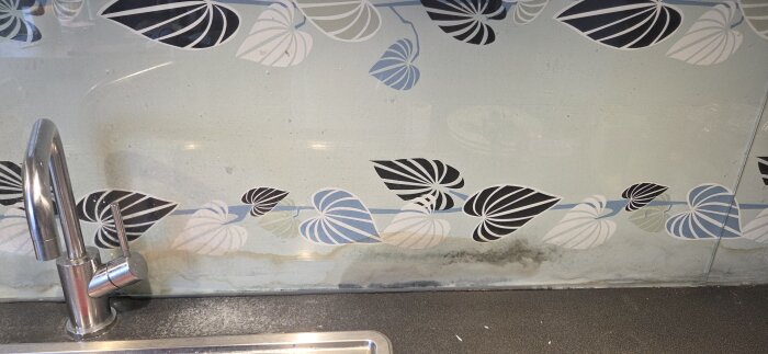 Close-up av köksblandare och stänkskydd med färgglad bladmönster. Mörka märken syns längs underkanten av glasskivan.