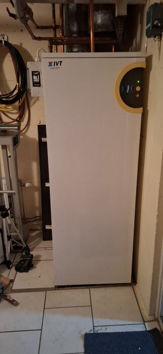 Bild på en vit IVT varmvattenberedare i ett källarutrymme, omgiven av kopparrör och verktyg på ett kaklat golv.