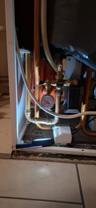 Bilden visar rörsystem med kopplingar och tryckmätare vid en värmepanna, med en flexibel slang ansluten, i en källare.