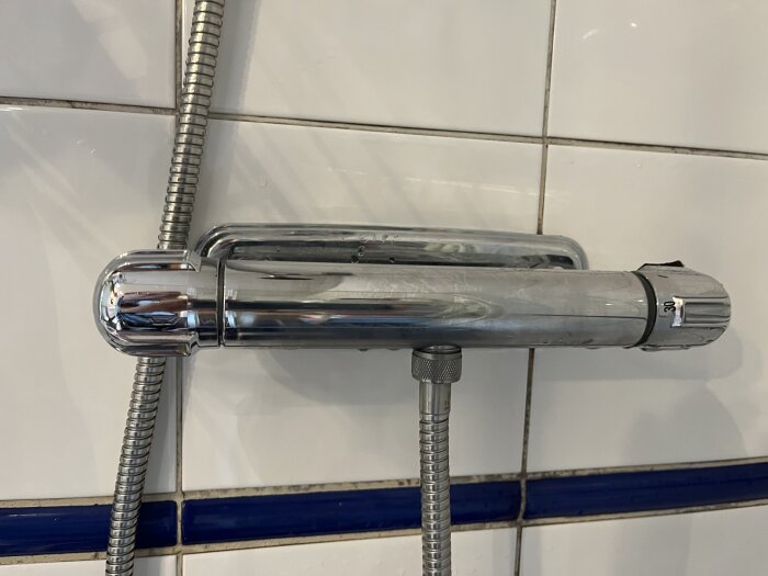 Duschblandare i krom, troligen av märket Mora Cera från 2003, installeras mot en vit kakelvägg med blå dekorlinje.