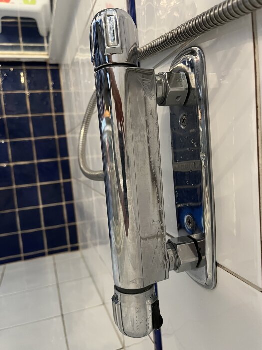 Kromfärgad duschblandare monterad på vitt kakel, med blått kakel i bakgrunden.