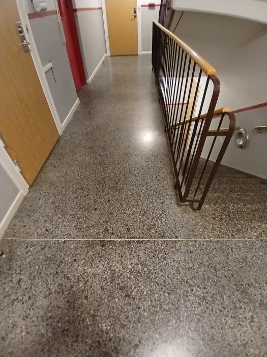 Trapphus med polerat sten- eller betonggolv, typiskt för byggnader från 1957, med träräcke och dörrar på sidan.