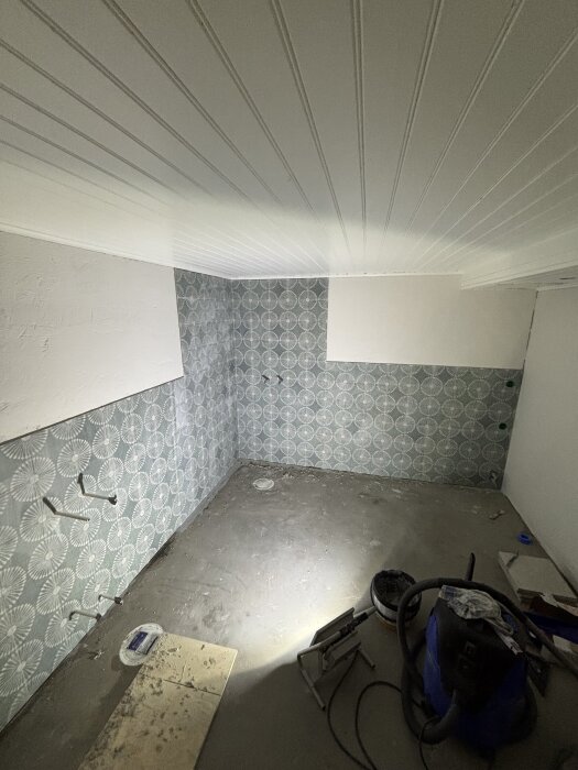 Nyfogade väggar i rum med mönstrade kakelplattor och vitmålat tak, golvberedning pågår med verktyg och rengöringsutrustning på golvet.