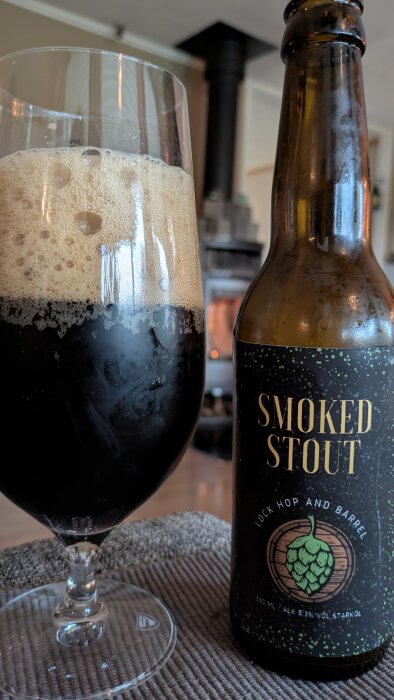 Flaska och glas med mörk Smoked Stout från Lock, Hop & Barrel, framför en tänd kamin.