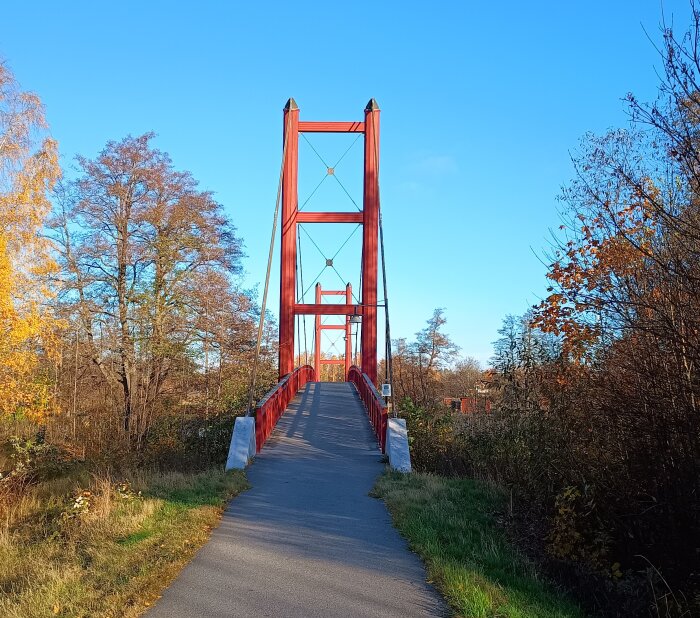 Röd bro i höstlandskap med blå himmel och träd med orange löv, fångar höstkänslan innan vinterns ankomst.