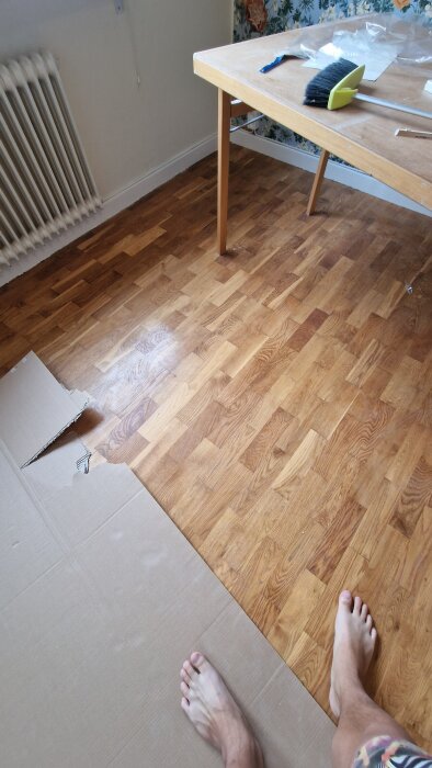 Barfota fötter på ett nylagt trägolv bredvid en bit kartong och ett bord med städmaterial.