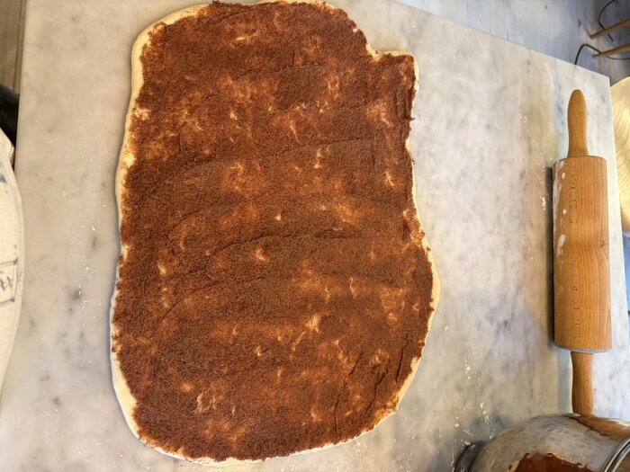 Utkavlad deg med kanel- och sockerblandning på en marmorbänk bredvid en kavel, för att baka kanelbullar.