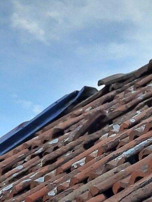 Närbild på ett tegelbelagt tak med ojämna rader och en plåt på högkantsavslutningen, delvis stöd av kloss nedtill.