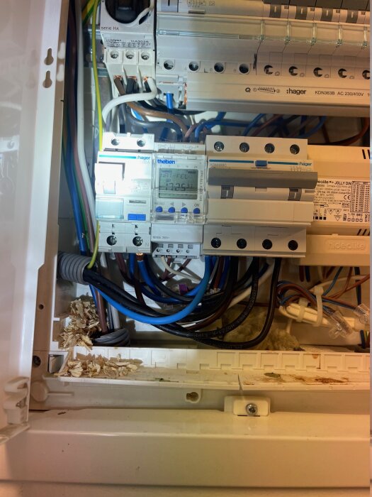 Elcentral med kablage och en Astro-ur i fokus, omgiven av trådar och isoleringsmaterial, med sågspån i botten av skåpet.