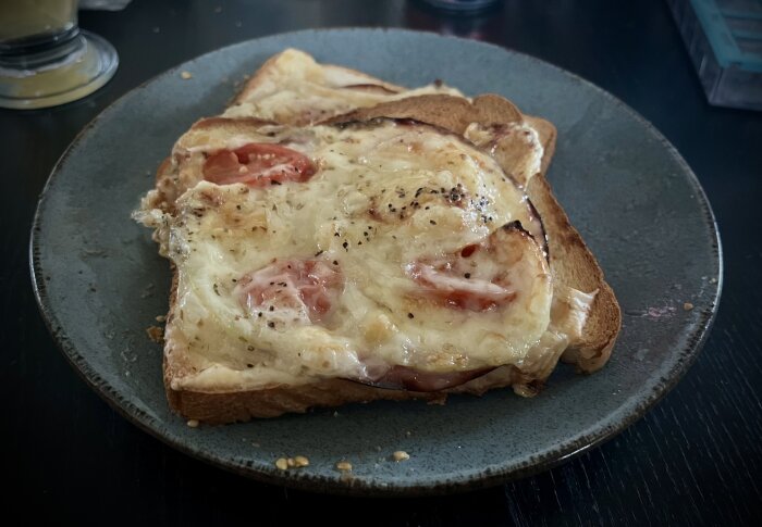 Varma mackor med smält ost, tomatskivor och svartpeppar på ett grått keramikfat.