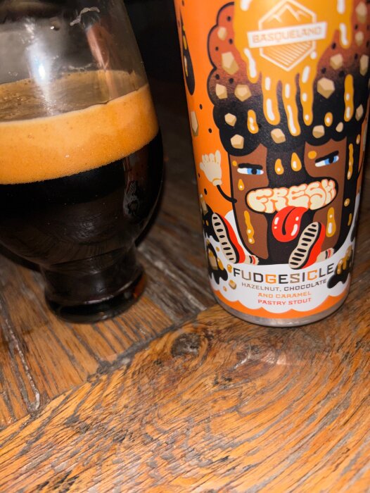 Ett glas mörk öl bredvid en färgglad burk med texten "Fudgesicle", en stout med hasselnöt, choklad och karamell.