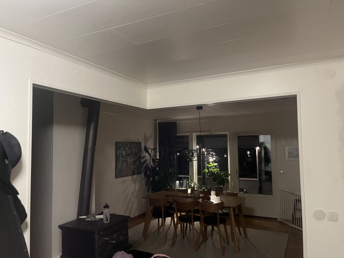 Öppen matsal med taklister, vita väggar och träbord med stolar. En kamin är placerad i hörnet, och taket har olika höjdnivåer.