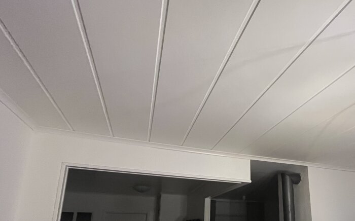 Vitt innertak med smala paneler och en öppning mot ett annat rum. Taklisten avslutas plötsligt vid en vägg.