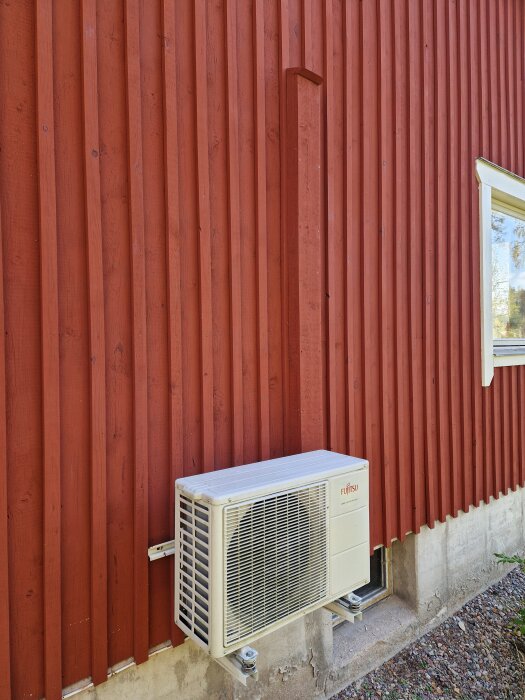Rödmålad fasad med tre brädor runt en vit luftkonditioneringsenhet, matchad med fasadens röda panel.