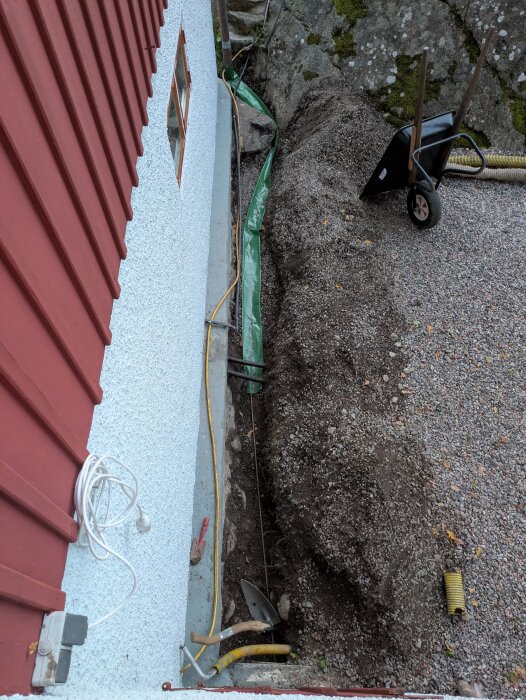 Markarbete intill en husgrund med en röd fasad. En skottkärra, kablar och en spade syns nära en grus- och jordhög.