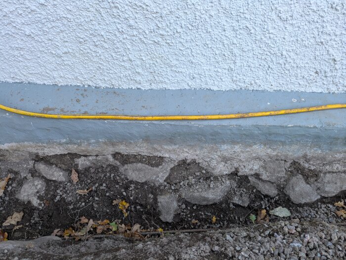 Vit vägg med gul kabel längs en grå sockel ovanför en grusig mark med stenar och löv.