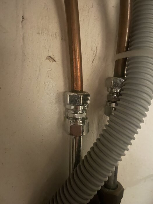 Två kopparrör med metallkopplingar, potentiellt ballofix och ett grått korrugerat plastslang i bakgrunden.