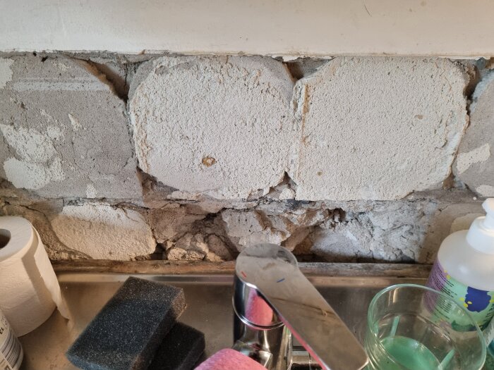Vägg över diskbänk med borttaget kakel, avslöjande ojämnt och skadat underlag. Rengöringsartiklar och kran syns i förgrunden.