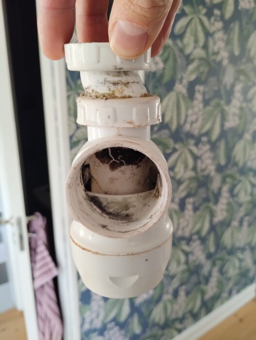 Gammalt, smutsigt vattenlås i plast med vinklad övre del, undersökt av hand, mot mönstrad tapetbakgrund.