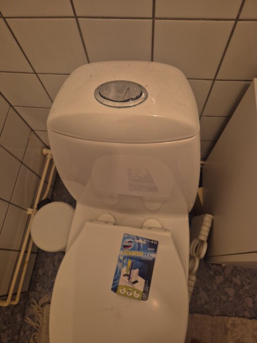 Toalett med ett paket WC-stICKS placerat på toalettlocket, klinkersvägg i bakgrunden.