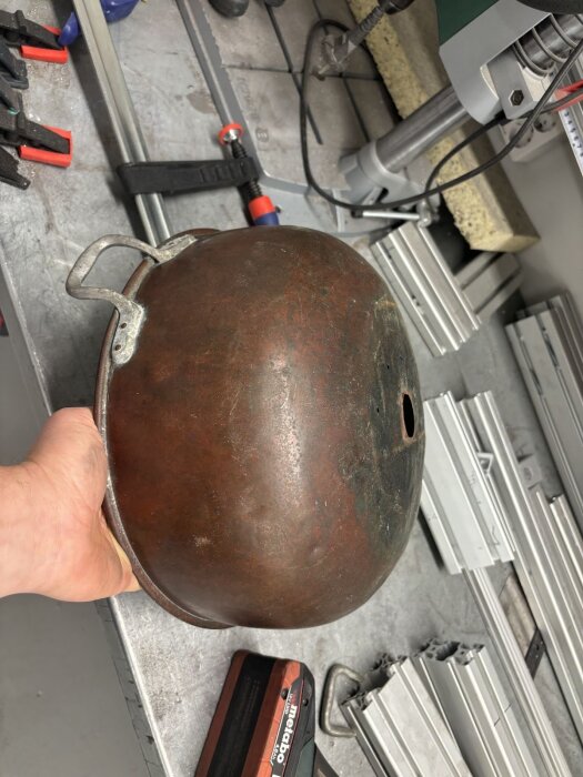 Ärgad kopparbunke med hål, omgjord med kulhammare för dekorativt bruk runt skorstensinstallation, visas på arbetsbänk.