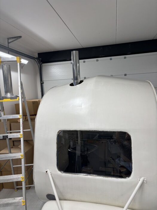 Skorstenen sticker upp genom taket på en vit vagn inomhus, med en stege och kartonger i bakgrunden.
