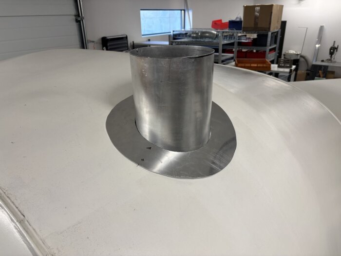 Aluminiumskorstensrör med metallkrage monterat på ett vitt tak i verkstadsmiljö, omgiven av olika byggmaterial och utrustning.