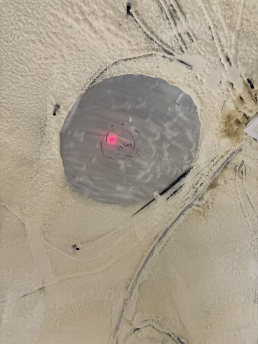Laserpekarens röda ljus markerar centrum för skorstenshålet genom ett tak, omgivet av struktur av skumisolering och takplåt.