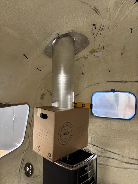 Aluminiumskorstensrör genom tak i vagn interiör, stöds av kartong ovanför kamin, med en nivå på kartongen för mätning och justering.