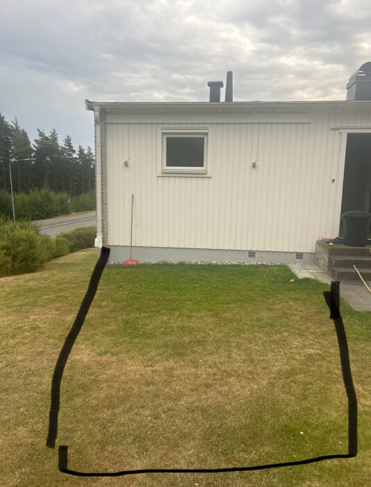 En vit byggnadsfasad med ett fönster, gräsmatta med markerad yta i svart, omgiven av träd i bakgrunden.