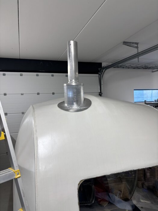 Aluminiumskorsten monterad på vit kupolformad yta i garage. Större rördel svetsad på plats. En del av stege synlig på sidan.