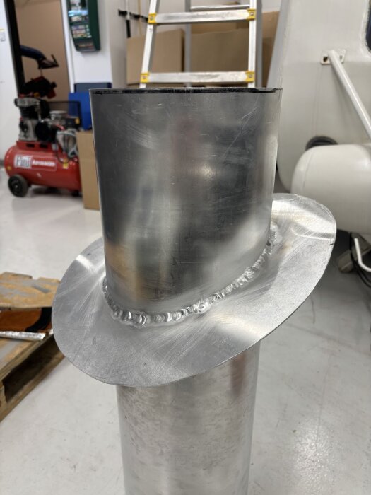 Aluminiumrör med svetsad krage, stående på ett verkstadsgolv med verktyg och stege i bakgrunden.